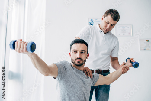 Chiropractor stretching arm of handsome patient with dumbbells in hospital