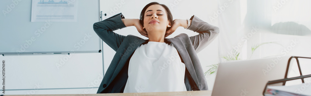panoramic shot of tired latin businesswoman relaxing with hands above head in office