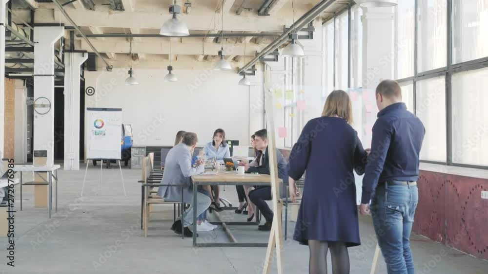 The business team at the big table holds a meeting. Nearby colleagues are drawing graphics on a glass board. Office life. Coworking