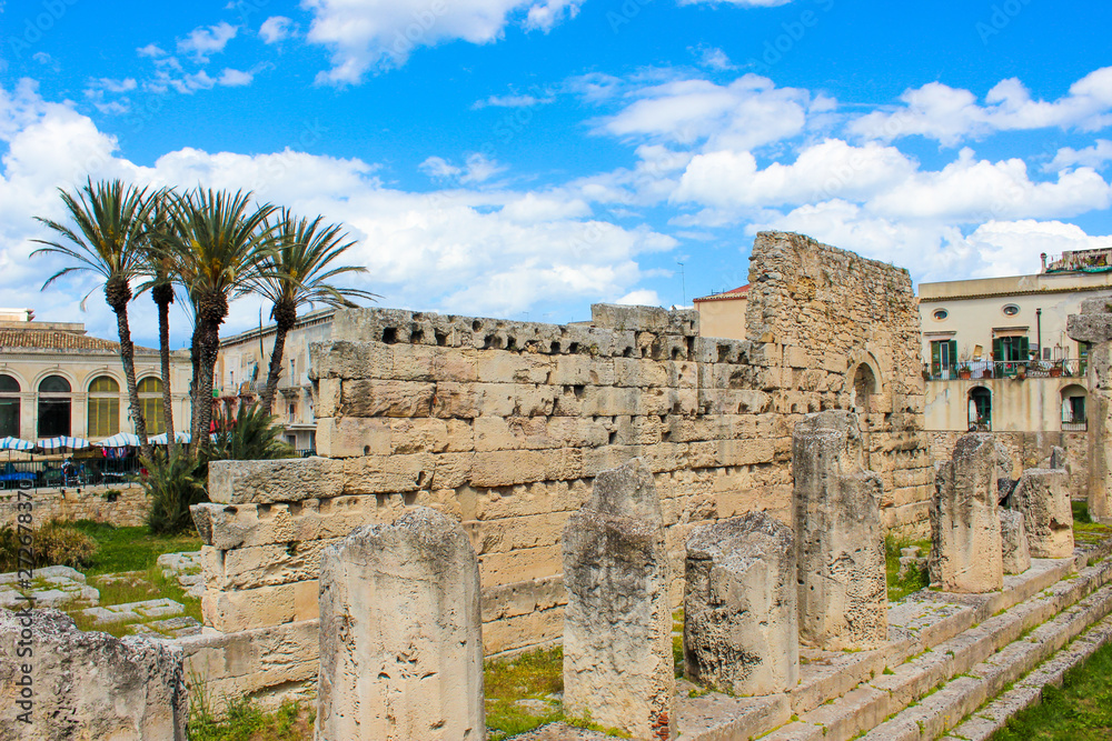 Significant ancient Greek ruins of the Temple of Apollo in Ortigia Island, Syracuse, Sicily, Italy. Colonnade remnants. Palm trees in background. Sunny day, blue sky. Popular tourist place