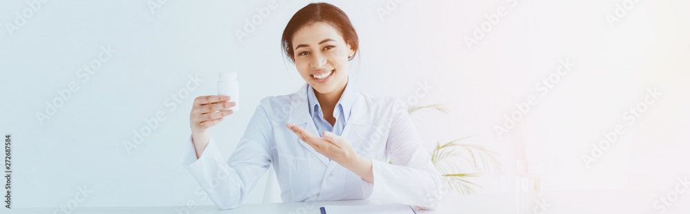 panoramic shot of smiling latin doctor pointing with hand at container with pills panoramic shot of smiling latin doctor pointing with hand at container with pills