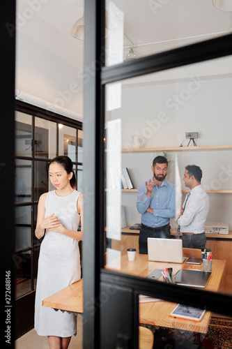 Fototapeta Naklejka Na Ścianę i Meble -  Asian young businesswoman standing near the entrance in board room and listening how two businessmen discussing her behind her back at office