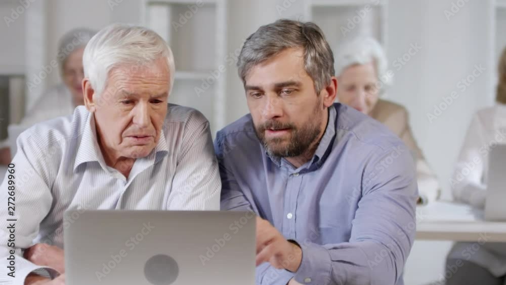 Tracking shot of middle-aged technology teacher helping senior man learning how to use laptop in computer class