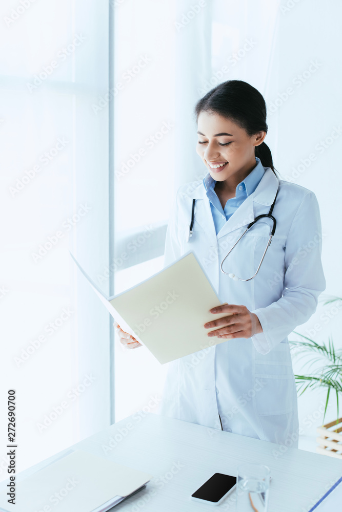 smiling latin doctor holding paper folder while standing by window in hospital smiling latin doctor holding paper folder while standing by window in hospital