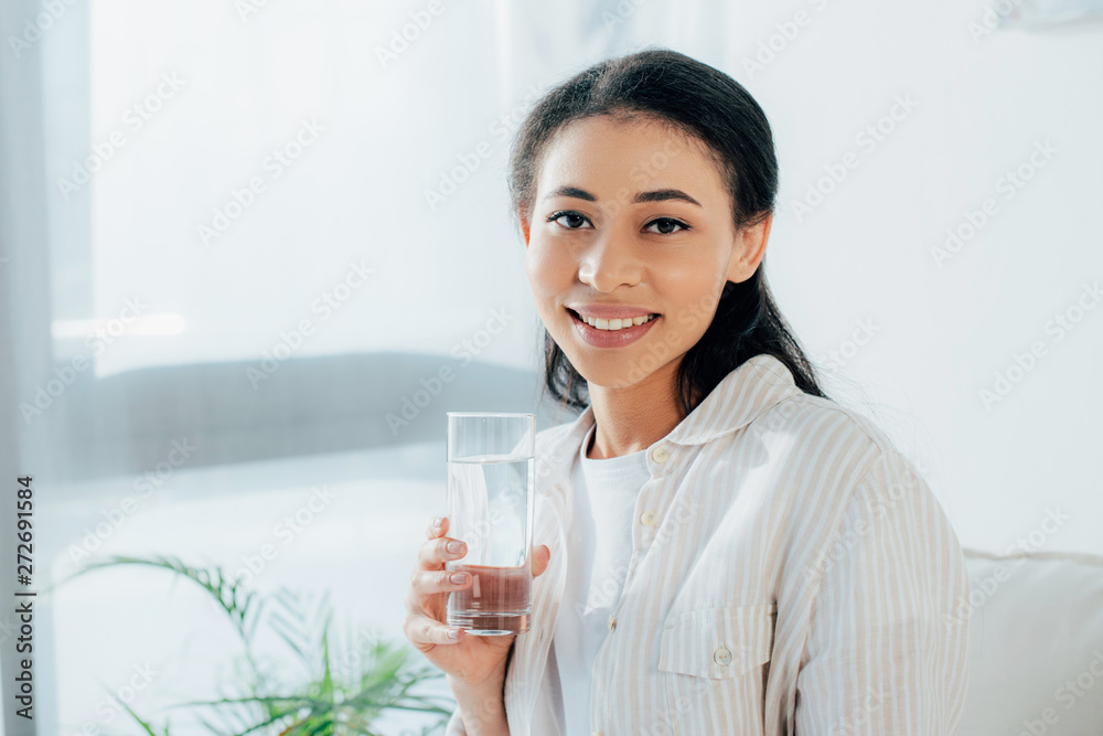 positive latin woman holding glass of water while smiling at camera positive latin woman holding glass of water while smiling at camera
