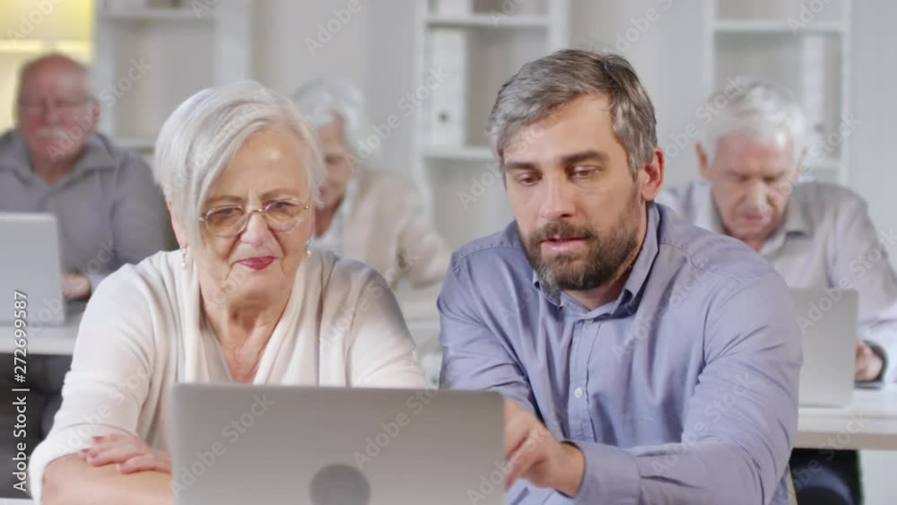 Tracking shot of male middle-aged technology teacher with beard helping elderly woman in glasses learning how to use laptop in computer class Tracking shot of male middle-aged technology teacher with beard helping elderly woman in glasses learning how to use laptop in computer class