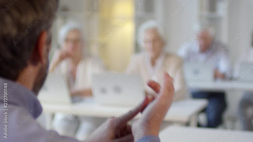 Handheld over the shoulder shot of unrecognizable male technology teacher gesturing and explaining how to use laptops to group of elderly people sitting at desks