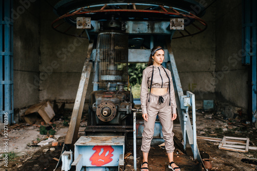 Beautiful young stylish girl posing at abandoned factory outdoor with an old broken engine. © benevolente