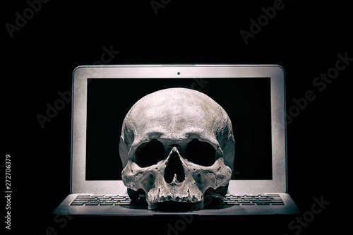 Cybercrime or hacking concept. Laptop in dark room under beam of light with a skull. Idea of virus or worm program cyber attack.