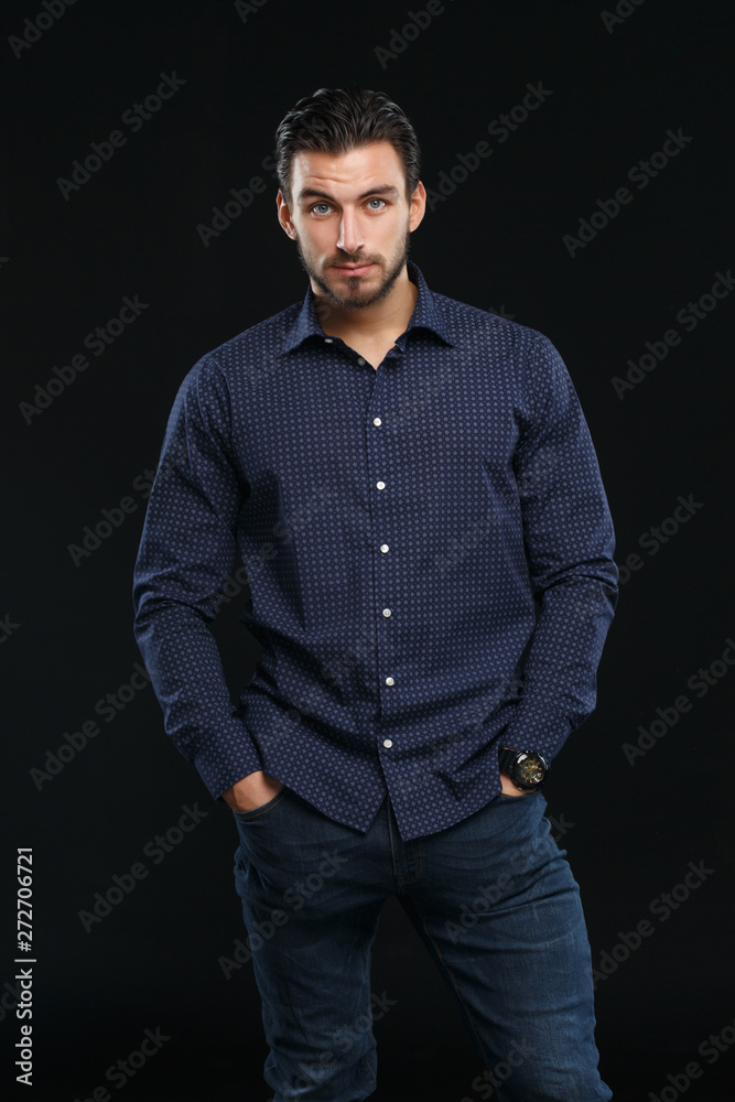 Portrait of a young bearded man in business casual clothes holding hands in his trouser pockets on a black background.