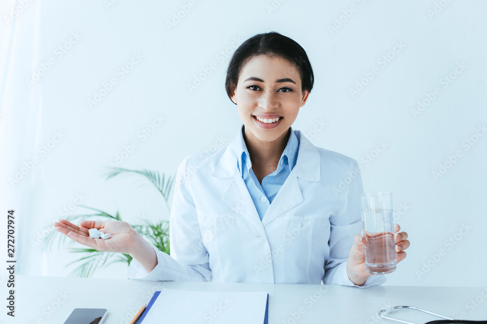 cheerful latin doctor with handful of pills and glass of water sitting at workplace and smiling at camera cheerful latin doctor with handful of pills and glass of water sitting at workplace and smiling at camera