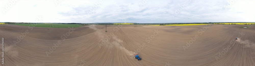 Aerial wide panoramic view on blue tractor pulling a plow, preparing a soil for seed sowing, tractor making dirt cloud. Landmark on meadow and rapeseed field