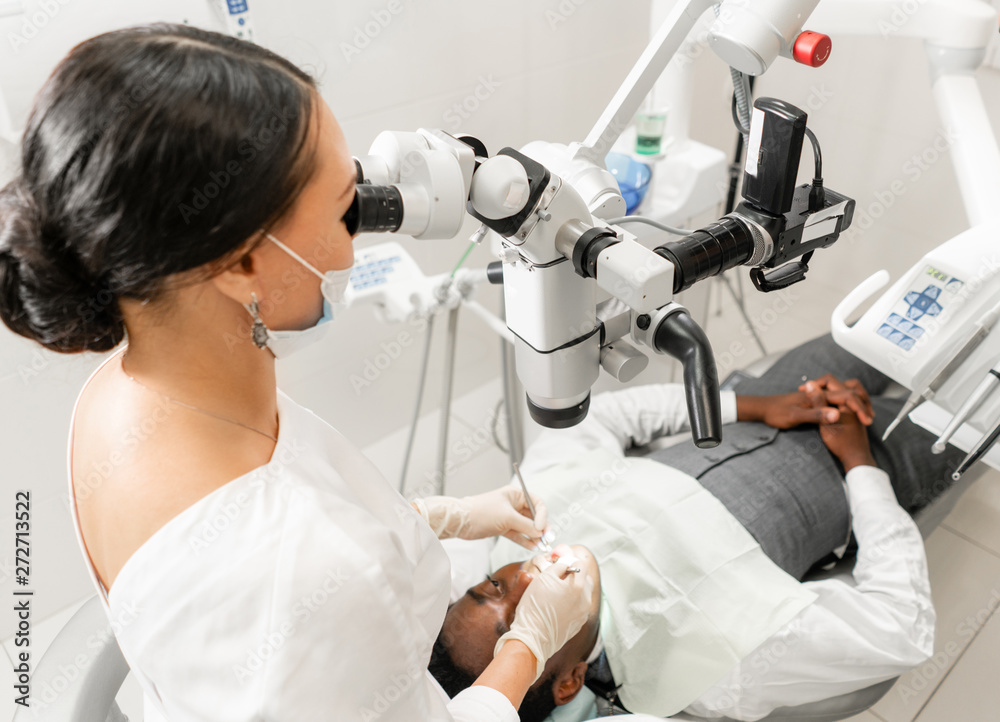 Young woman dentist treating root canals using microscope in the dental clinic. Man patient lying on dentist chair with open mouth. Medicine, dentistry and health care concept. Dental equipment Young woman dentist treating root canals using microscope in the dental clinic. Man patient lying on dentist chair with open mouth. Medicine, dentistry and health care concept. Dental equipment