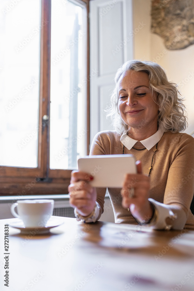 Portrait of smiling mature businesswoman sitting at table with cup of coffee looking at cell phone Portrait of smiling mature businesswoman sitting at table with cup of coffee looking at cell phone