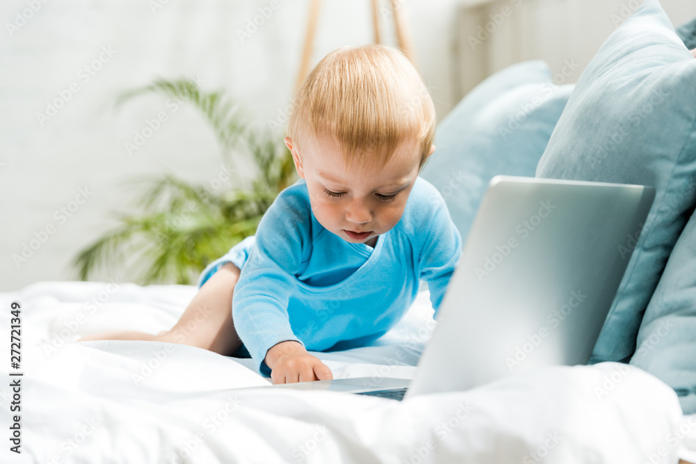 selective focus of cute toddler kid sitting on bed near laptop at home
