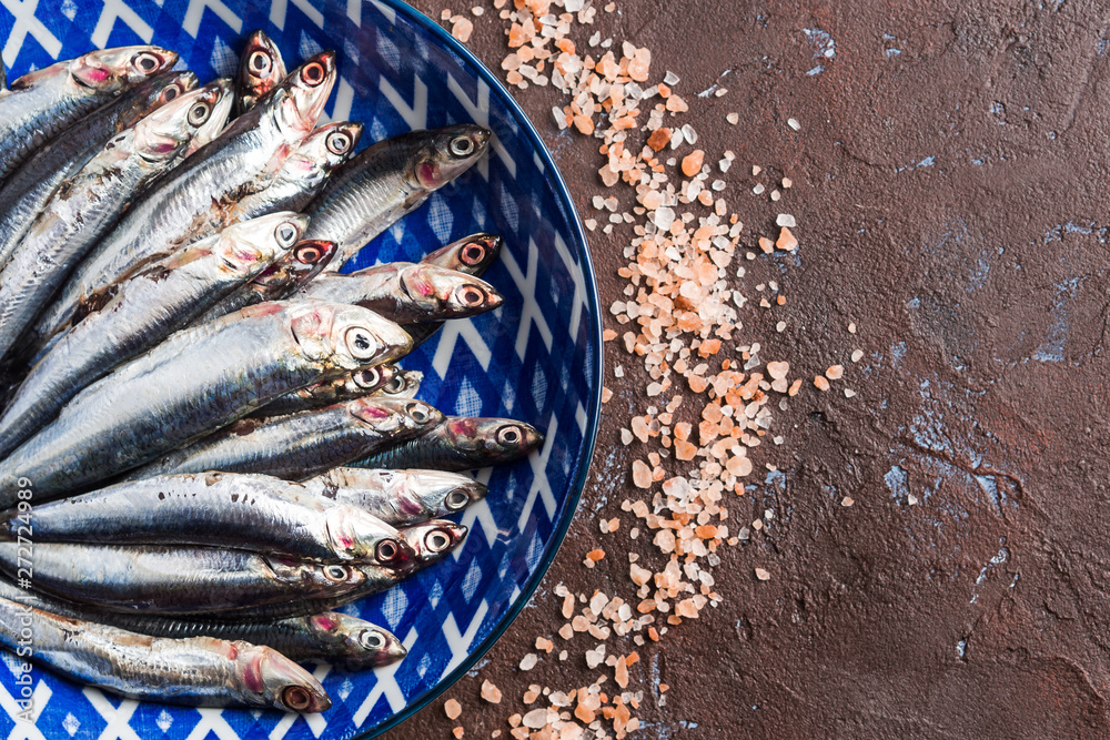 Blue fish. Fresh anchovies in a dish with pink salt on dark brown background. Healthy protein meal. Local seafood, Italy Blue fish. Fresh anchovies in a dish with pink salt on dark brown background. Healthy protein meal. Local seafood, Italy