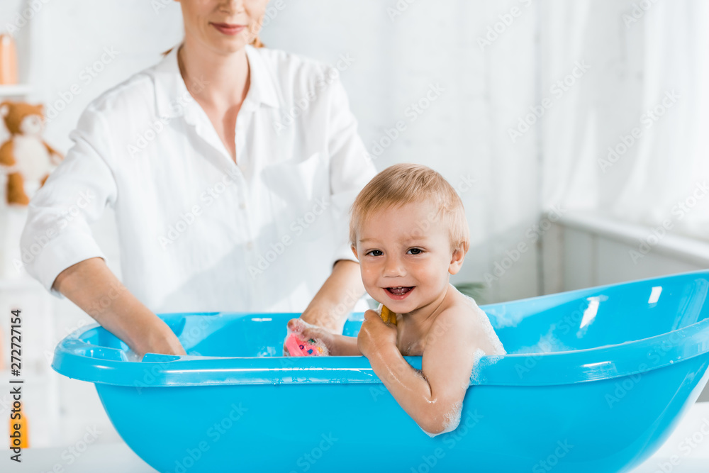 selective focus of cute smiling toddler boy in baby bathtub near mother selective focus of cute smiling toddler boy in baby bathtub near mother