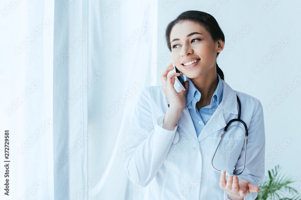 pretty young doctor talking on smartphone while standing by window in hospital
