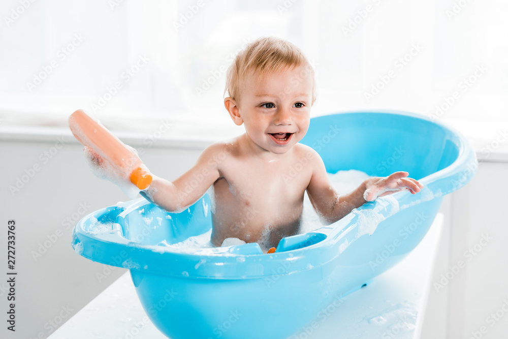 happy toddler kid smiling while taking bath in blue baby bathtub and holding bottle with shampoo happy toddler kid smiling while taking bath in blue baby bathtub and holding bottle with shampoo