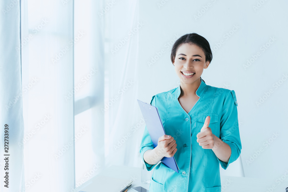 happy latin doctor showing thumb up and smiling at camera while standing by window in hospital happy latin doctor showing thumb up and smiling at camera while standing by window in hospital