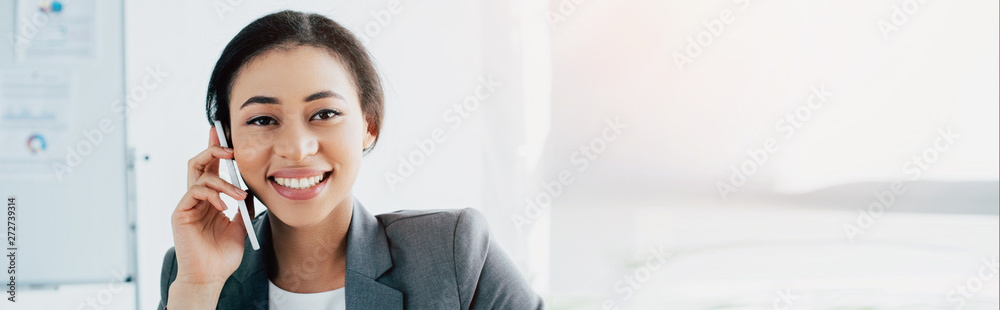 panoramic shot of cheerful latin businesswoman talking on smartphone and smiling at camera panoramic shot of cheerful latin businesswoman talking on smartphone and smiling at camera