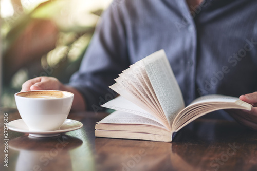 Closeup image of a woman holding and reading a book while drinking coffee on wooden table