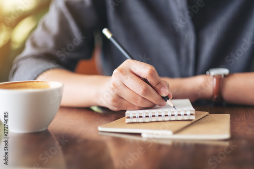 Closeup image of a woman's hand writing on blank notebook with coffee cup on table in cafe