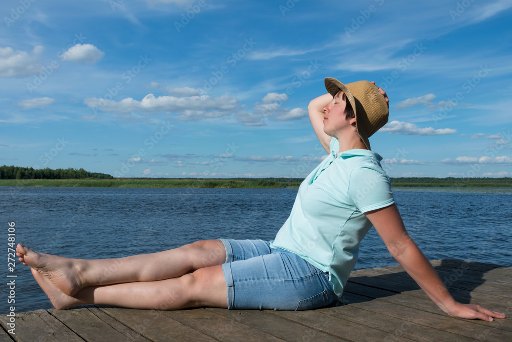 girl in a hat sunbathes on a pier by the lake on a hot sunny day