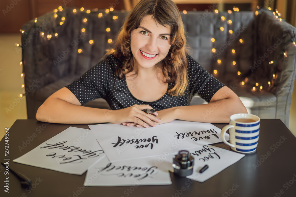 Fototapeta premium Love yourself. Calligrapher Young Woman writes phrase on white paper. Inscribing ornamental decorated letters. Calligraphy, graphic design, lettering, handwriting, creation concept