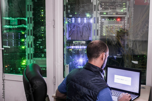 An employee of a telecommunications company works in a data center. A man with a laptop is sitting in a dark server room.