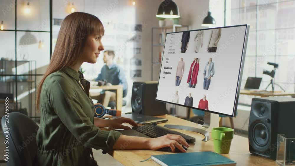 Beautiful Brunette Female Scrolls Through Online Fashion Store on Her Personal Computer with Big Display. She Works in a Cool Office Loft. Other Male Creative Colleague Walks in the Background.