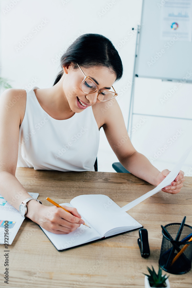 Obraz premium smiling latin businesswoman writing in notebook while sitting at workplace in office