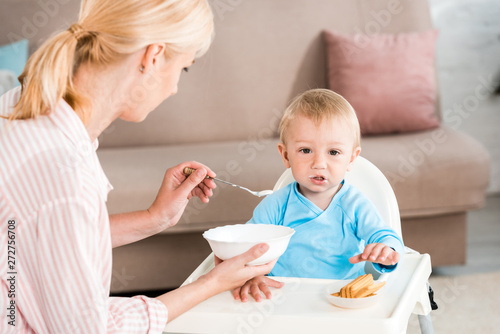 selective focus of blonde mother holding bowl and feeding cute toddler son at home
