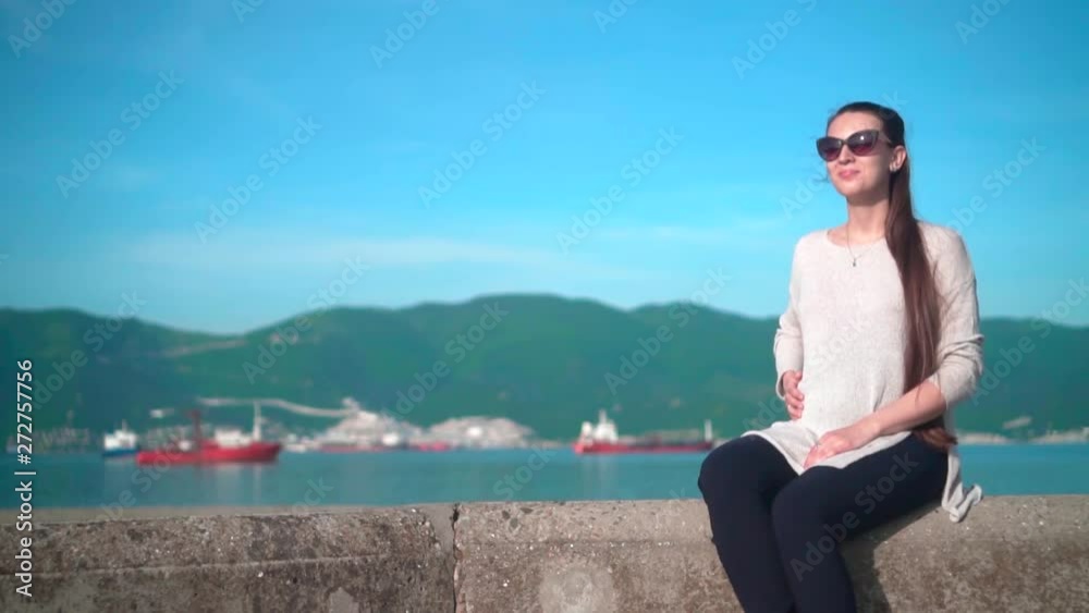 Young pregnant charming girl in sunglasses in clear weather in the summer. The girl sits on the embankment near the sea on the background of mountains and ships. Young pregnant charming girl in sunglasses in clear weather in the summer. The girl sits on the embankment near the sea on the background of mountains and ships.