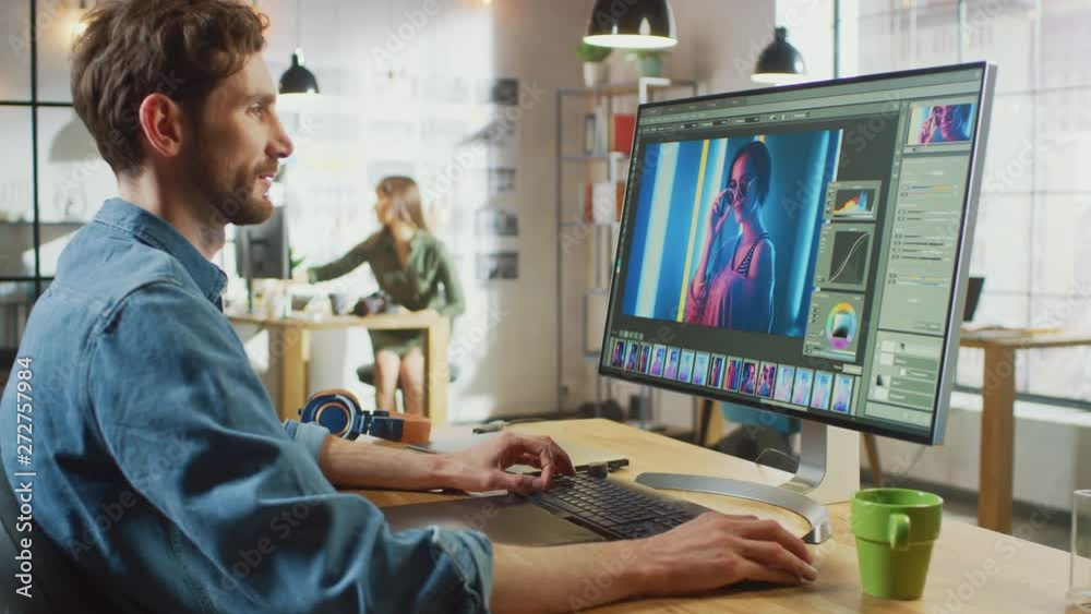 Male Digital Editor in Jeans Shirt Works in Picture Editing Software on His Personal Computer with Big Display. He Works in Cool Office Loft. Other Female Creative Colleague Walks in the Background. Male Digital Editor in Jeans Shirt Works in Picture Editing Software on His Personal Computer with Big Display. He Works in Cool Office Loft. Other Female Creative Colleague Walks in the Background.