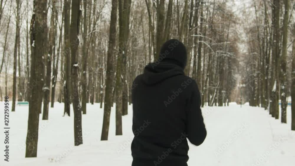 Steadicam shot rear view of a young guy in black sportswear with a hood performing a hand-held workout in a winter snow-covered city Park. The concept of hardening fighter, puncher, lover of martial Steadicam shot rear view of a young guy in black sportswear with a hood performing a hand-held workout in a winter snow-covered city Park. The concept of hardening fighter, puncher, lover of martial
