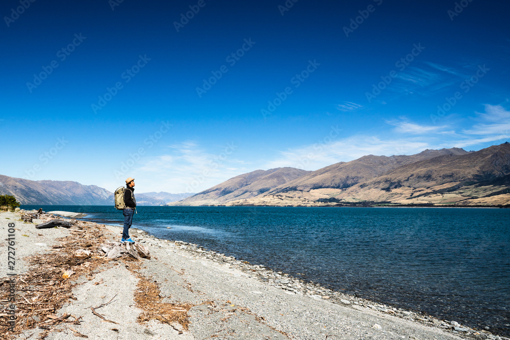 Obraz premium Tourist admires the view while stand next to Lake wanaka Boundary Creek with background of blue sky and rocky mountain with copy space.