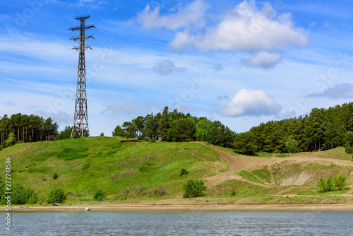 View from the river to the wooded shore and high-voltage tower. Blue sky with clouds on summer day