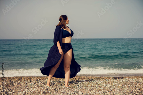 Attractive busty curvy woman in a blue swimsuit resting on the beach. Stylish accessories, fringe, fashion for plus size, beautuful sea. Bodypositive, natural authentic beauty, resort, summer vacation photo