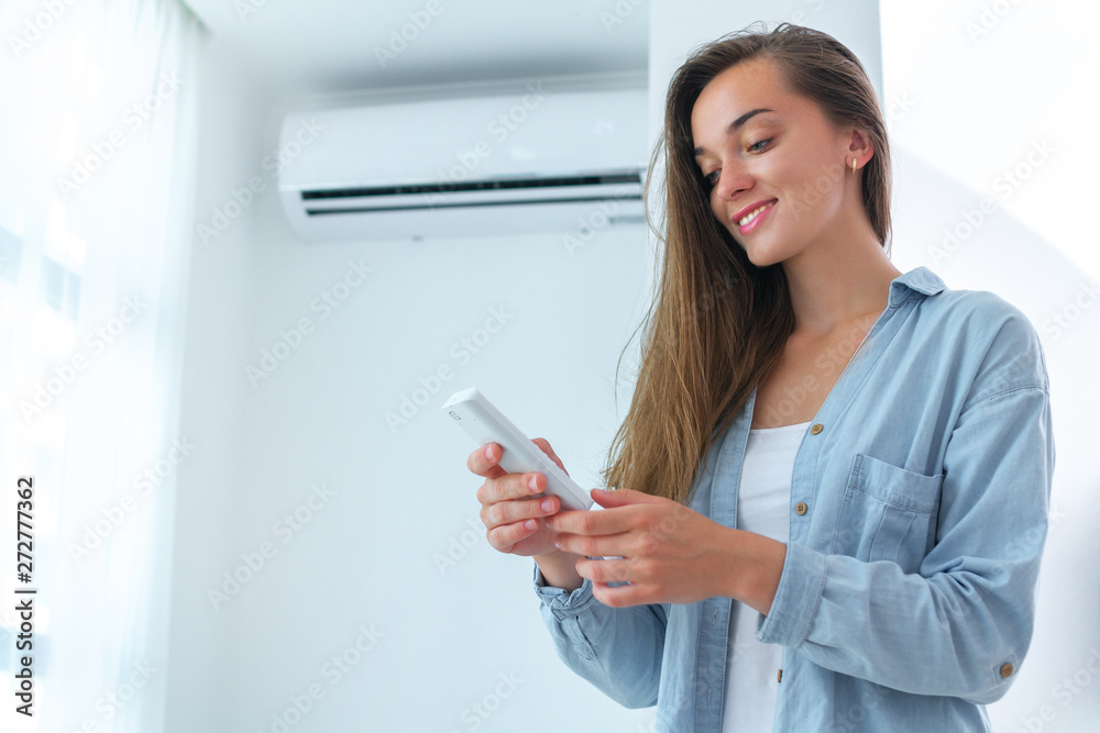 Young, happy, smile woman adjusts the temperature of the air conditioner using the remote control in room at home Young, happy, smile woman adjusts the temperature of the air conditioner using the remote control in room at home