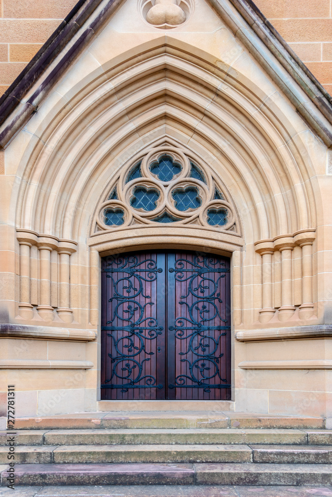 Red and brown church entrance door with ornate wrought iron recessed into an ornate  textured sandstone pointed archway
