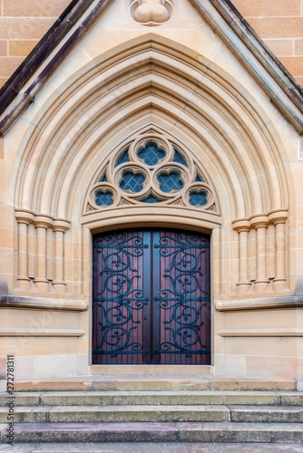 Red and brown church entrance door with ornate wrought iron recessed into an ornate textured sandstone pointed archway