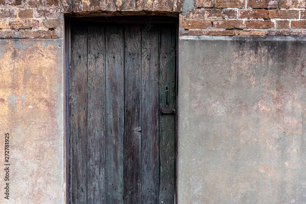 Close up of an old textured and weathered dark grey and brown wooden entrance door with stained concrete and brick wall