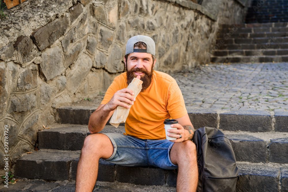 Fastfood break. Bearded man eating hot dog for meal break. Hipster tourist relaxing on stairs during rest break. Caucasian guy traveler having coffee break on urban background