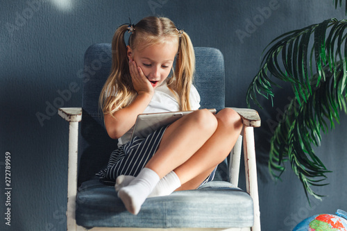 little blondy girl sitting on a chair and doing homework for school, researching information on the tablet.