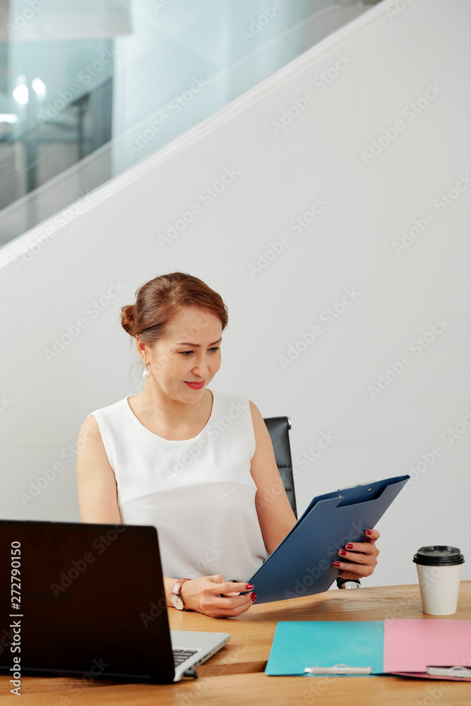 Confident manager sitting at the table in front of laptop computer and looking at blue folder in her hands Confident manager sitting at the table in front of laptop computer and looking at blue folder in her hands