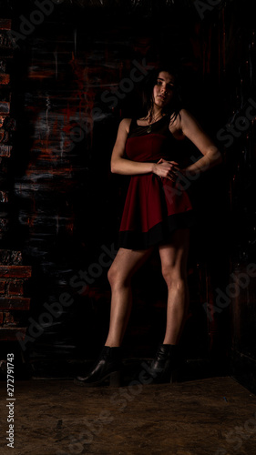 Pretty young sexy model female with dark hair in amazing long red dress and black shoes posing in dark studio