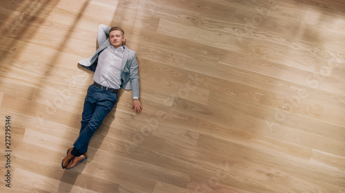 Young Man is Lying on a Wooden Flooring in an Apartment. He's Wearing a Jacket and White Shirt. Cozy Living Room with Modern Minimalistic Interior and Wooden Parquet. Top View Camera Shot. © Gorodenkoff