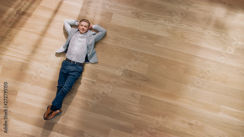 Young Man is Lying on a Wooden Flooring in an Apartment. He's Wearing a Jacket and White Shirt. Cozy Living Room with Modern Minimalistic Interior and Wooden Parquet. Top View Camera Shot.