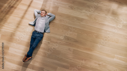 Young Man is Lying on a Wooden Flooring in an Apartment. He's Wearing a Jacket and White Shirt. Cozy Living Room with Modern Minimalistic Interior and Wooden Parquet. Top View Camera Shot.